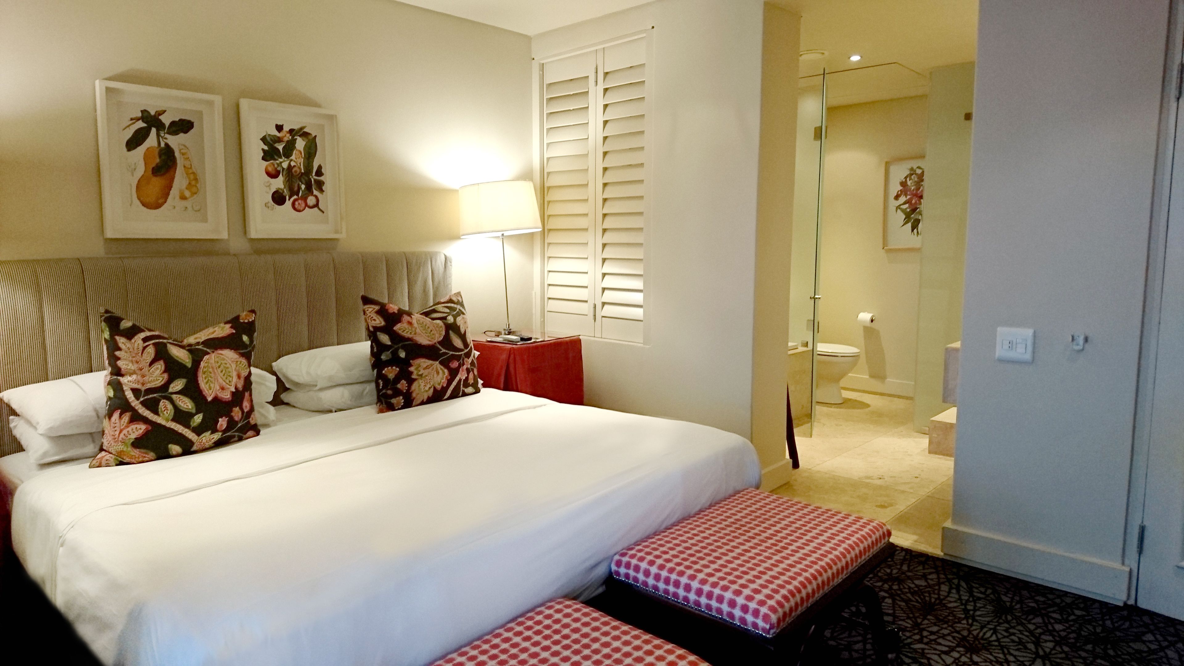 A hotel room with a large bed, two pillows, a bedside table, a lamp, and a window with shutters.