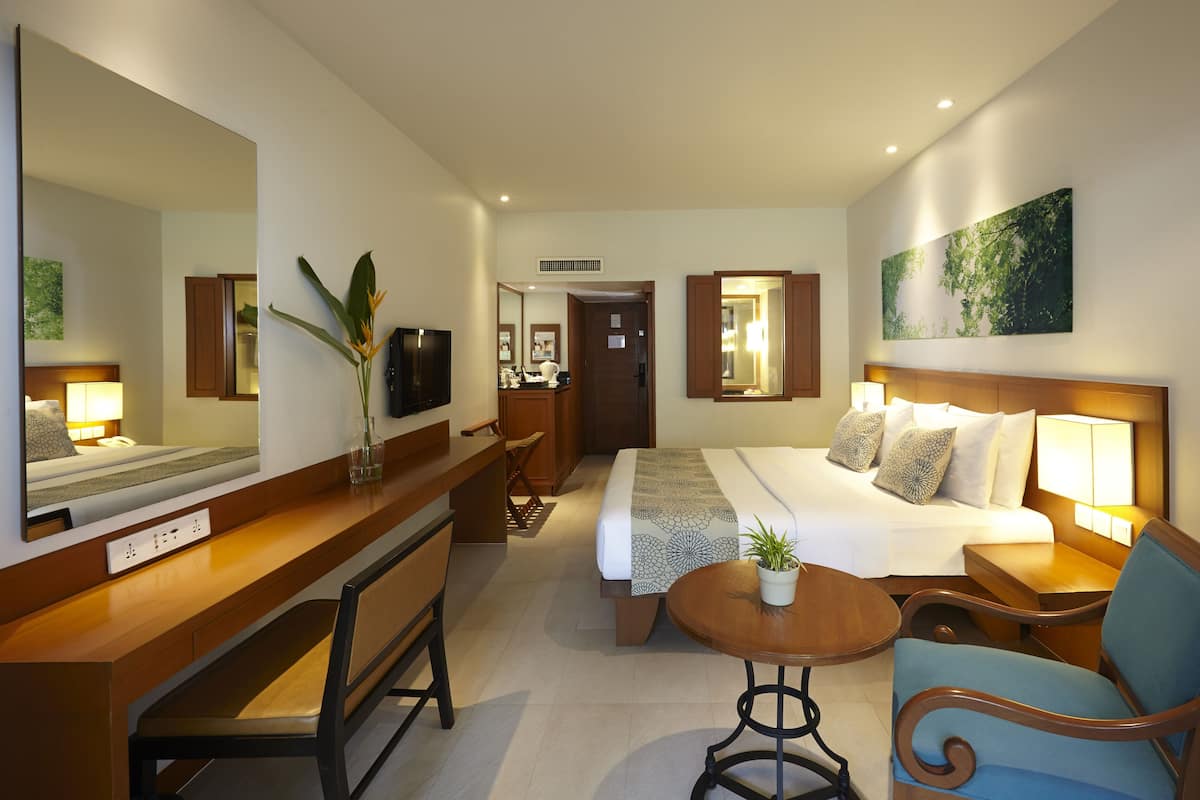 Une chambre d’hôtel comprenant un lit, des tables de chevet, un bureau, une chaise, une petite table, un miroir, un téléviseur et une plante en pot.