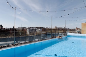 Una piscina en la azotea con agua azul cristalina, rodeada por una cerca de metal y luces colgantes.