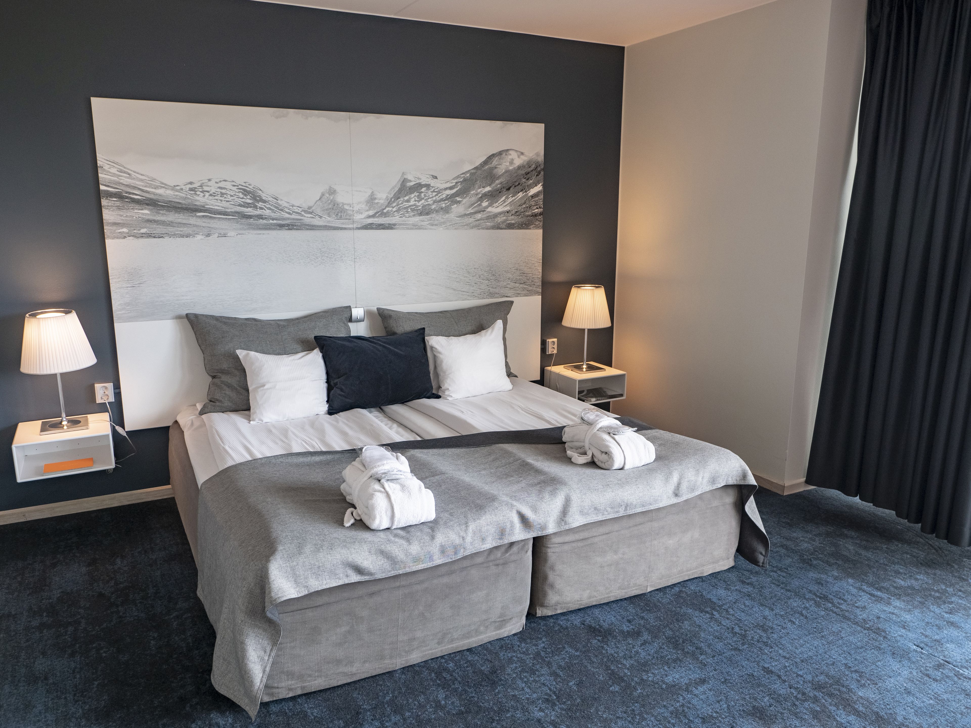 Una habitación de hotel moderna con una cama grande, dos lámparas de noche, un cabecero con una pintura de un paisaje y vistas a un lago de montaña.