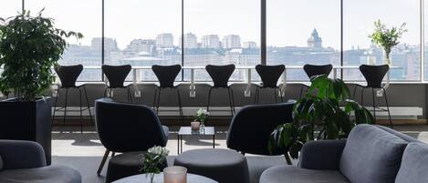 A modern lounge area with a city view, featuring sofas, chairs, and a round table with a vase and candle.