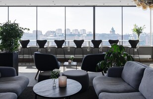 Un área de estar moderna con vistas a la ciudad, que incluye sofás, sillas y una mesa redonda con un florero y una vela.