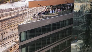 A modern building with a glass facade, a rooftop terrace with people, and a railway track below.