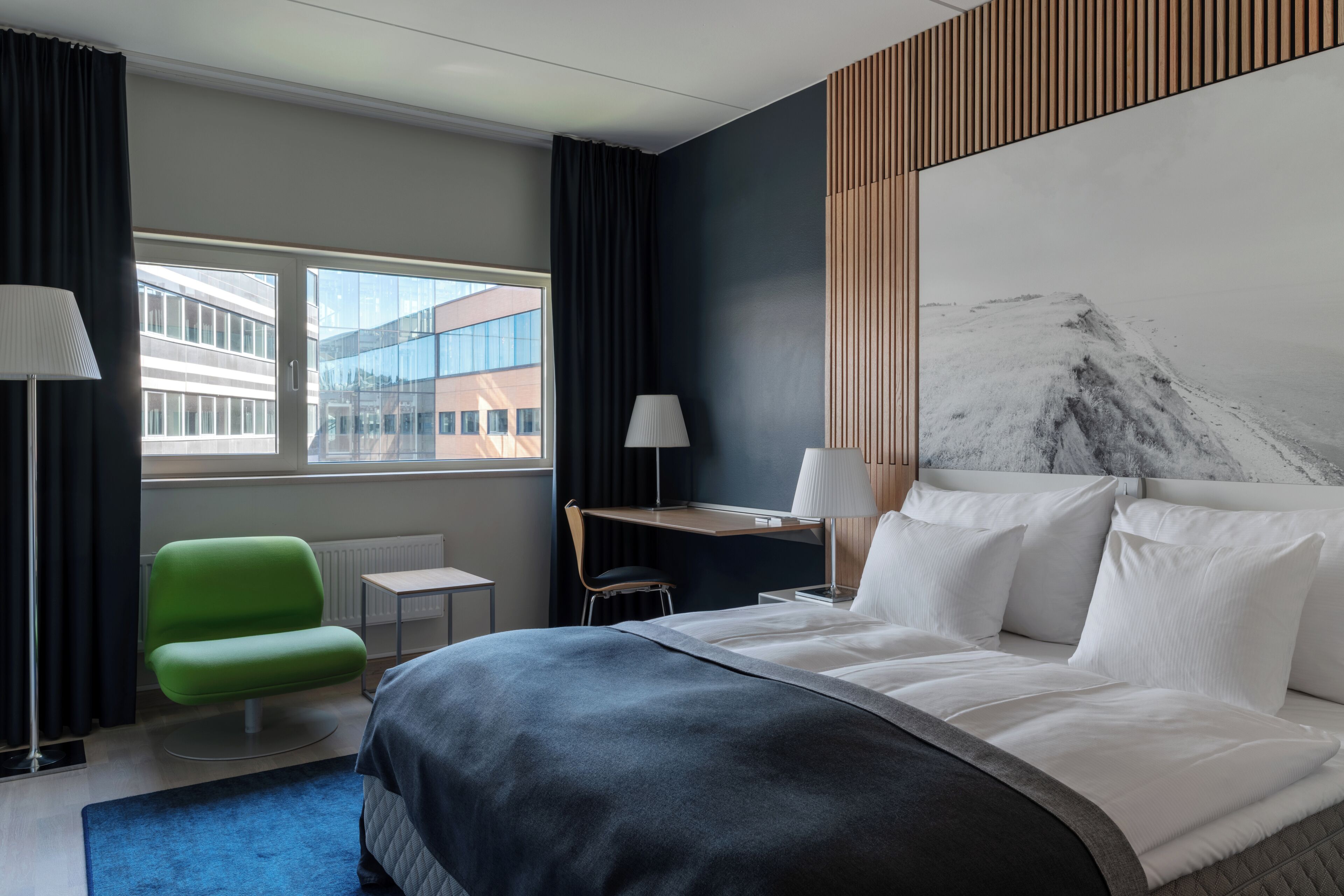 A modern hotel room with a large bed, a desk, a chair, and a view of a building through the window.