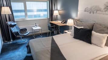 Habitación de hotel con cama, escritorio, silla, lámpara y vistas al exterior.