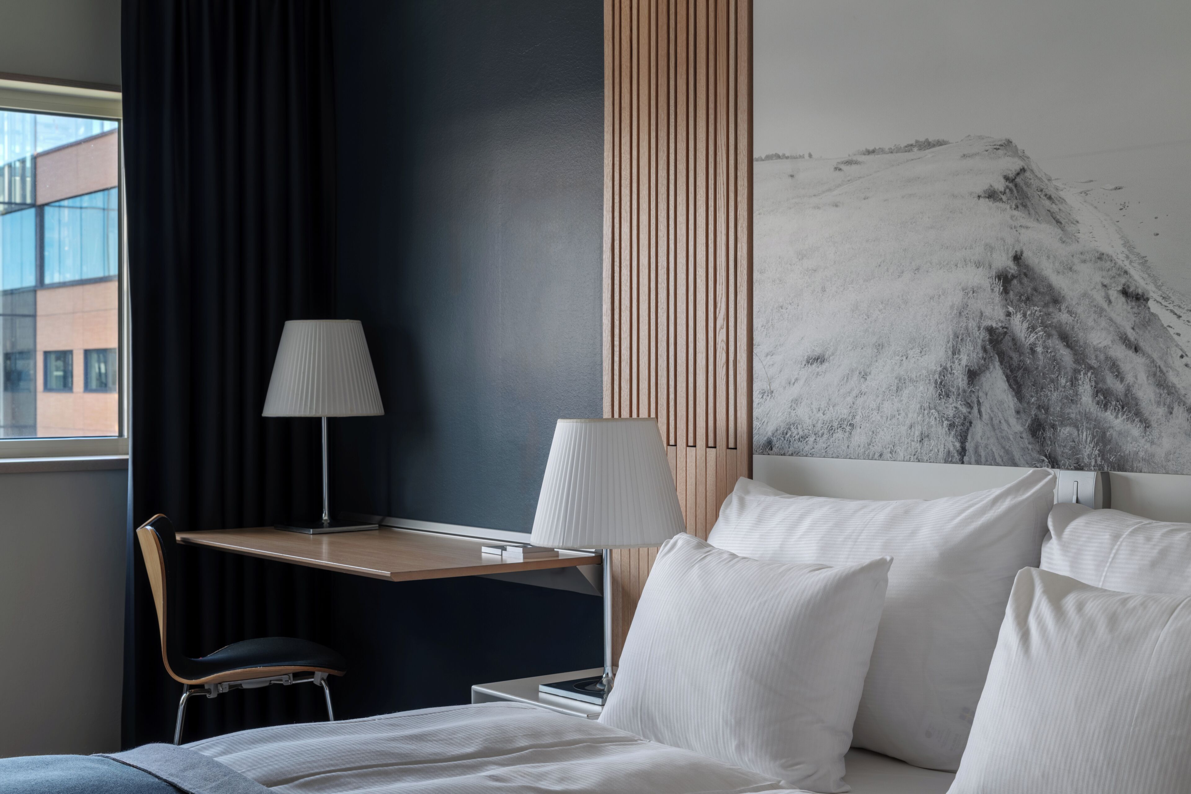 A hotel room with a bed, a desk with a chair, a lamp, and a large abstract painting on the wall.