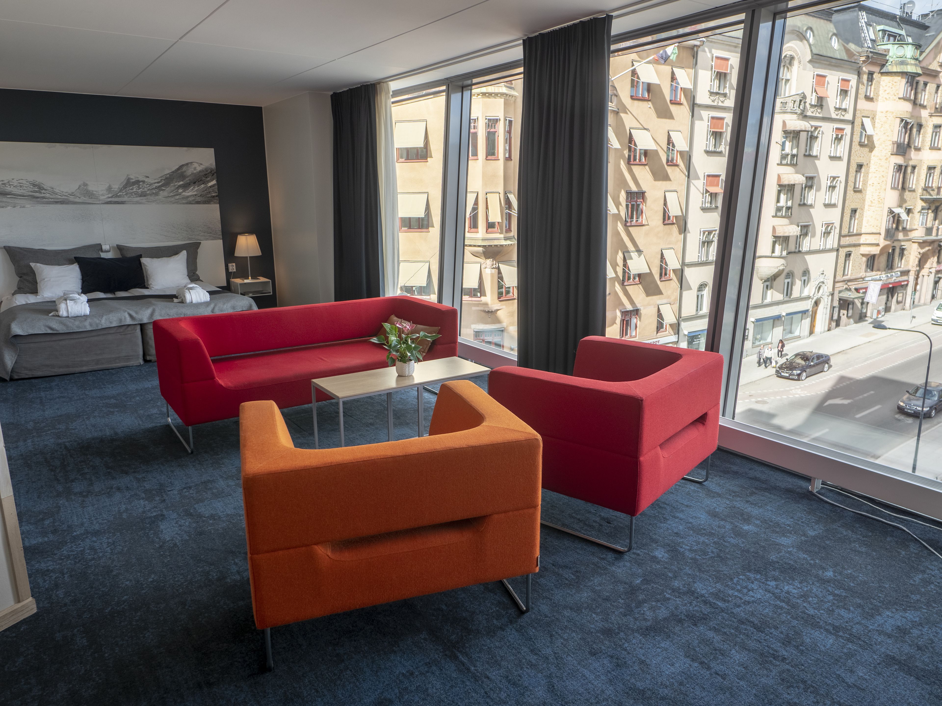 Een hotelkamer met een bed, een rode bank, een oranje fauteuil, een klein tafeltje met een plant en grote ramen met uitzicht op de stad.