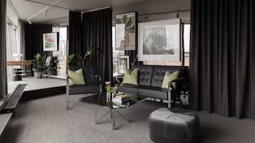Una sala de estar moderna con una mesa de centro de vidrio, un sofá de cuero con cojines verdes y un reposapiés gris. Hay cortinas negras, un cuadro enmarcado en la pared y una ventana con vistas a edificios.