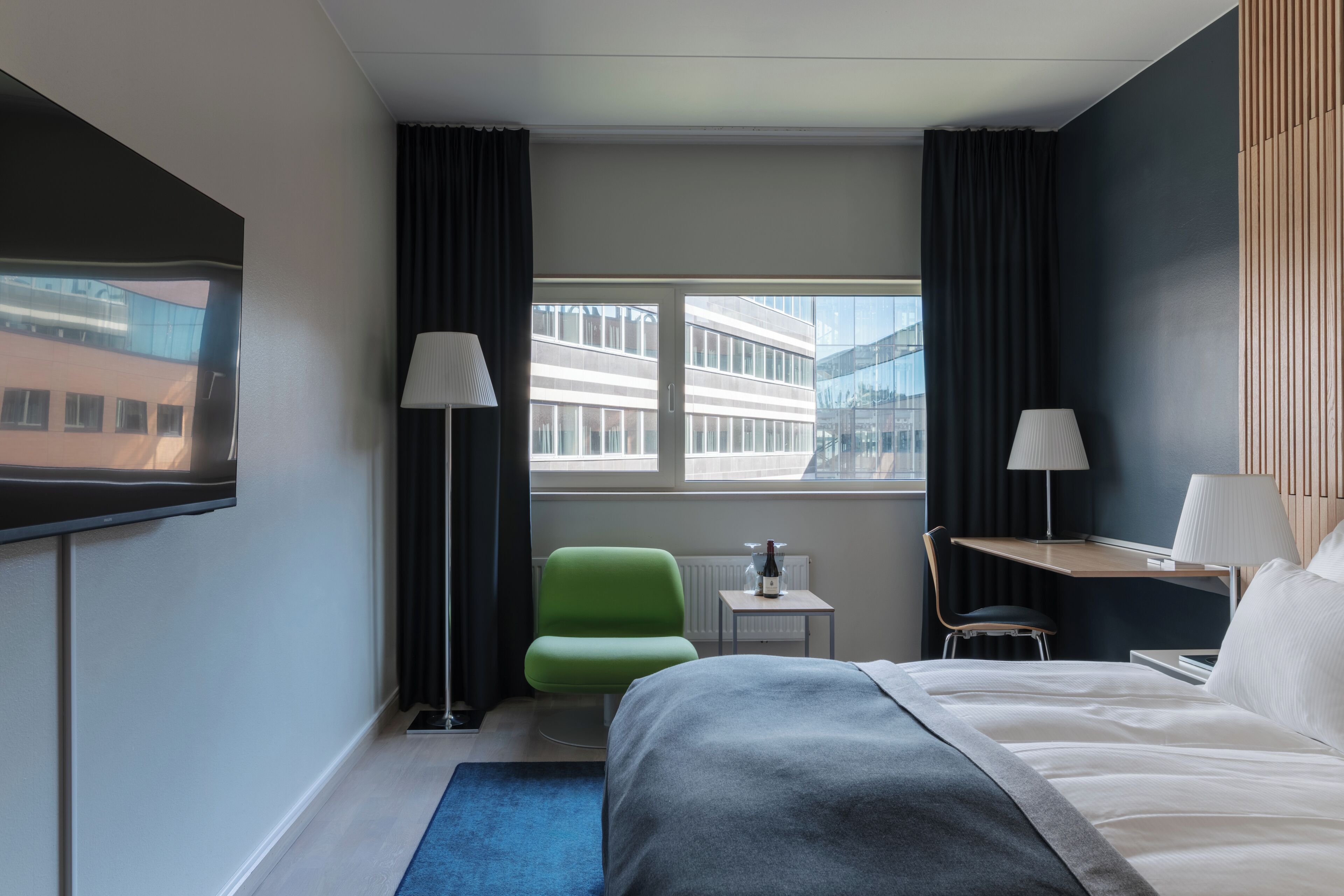Habitación de hotel con cama, escritorio, silla, lámpara, ventana y vista a un edificio.