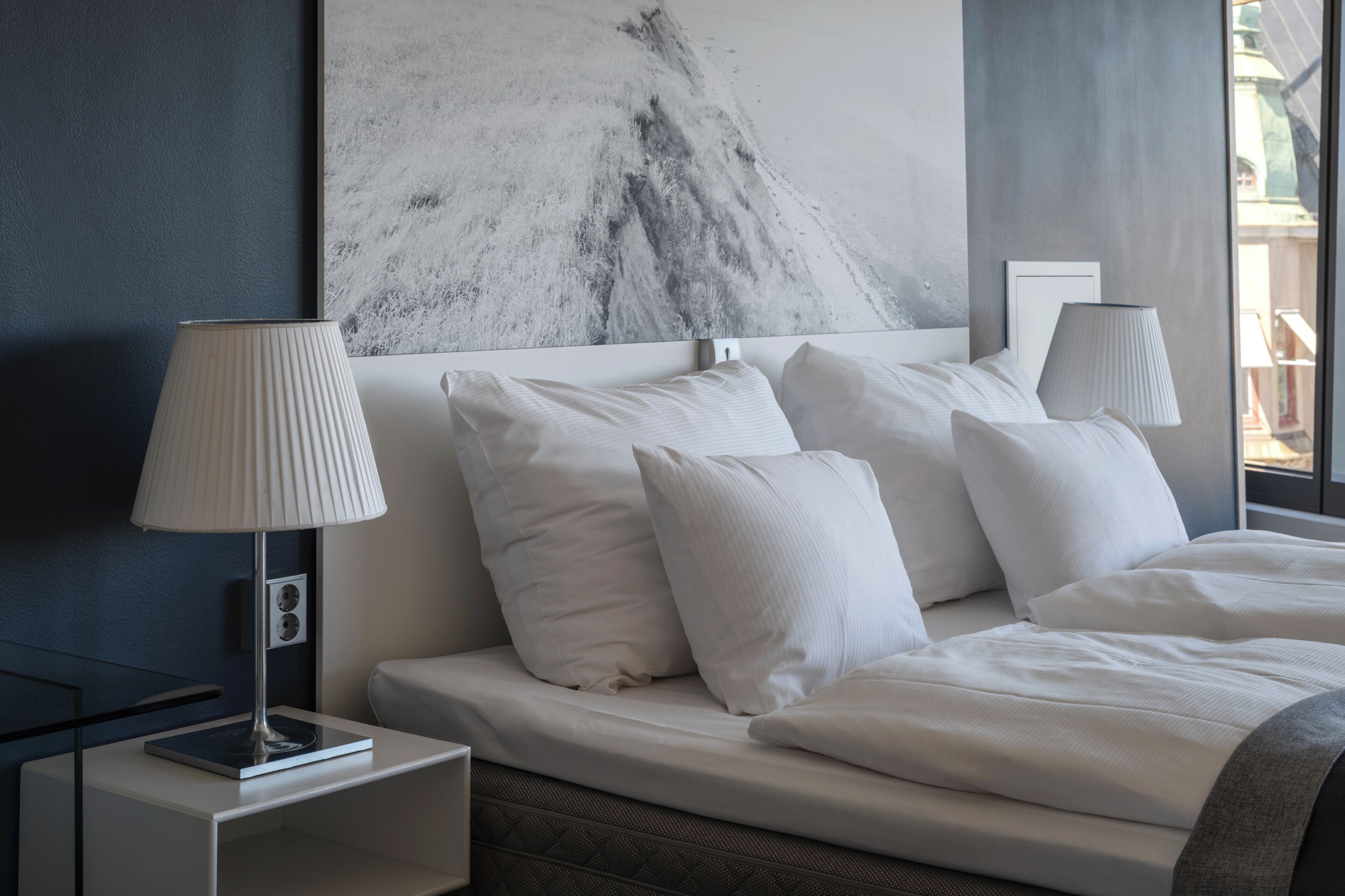 A modern hotel room with a large bed, white bedding, a marble artwork on the wall, and a nightstand with a lamp.