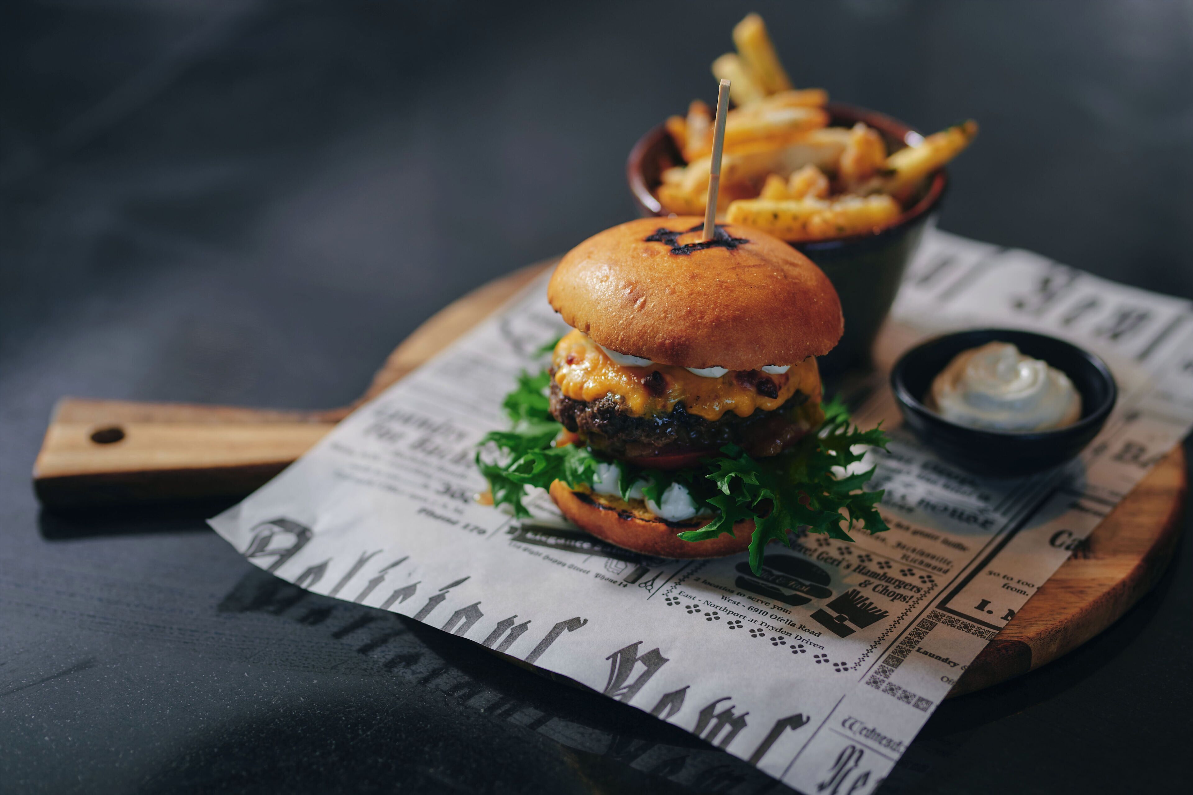 Burger med pommes frites og en lille skål sauce på en træplade med en avis under.
