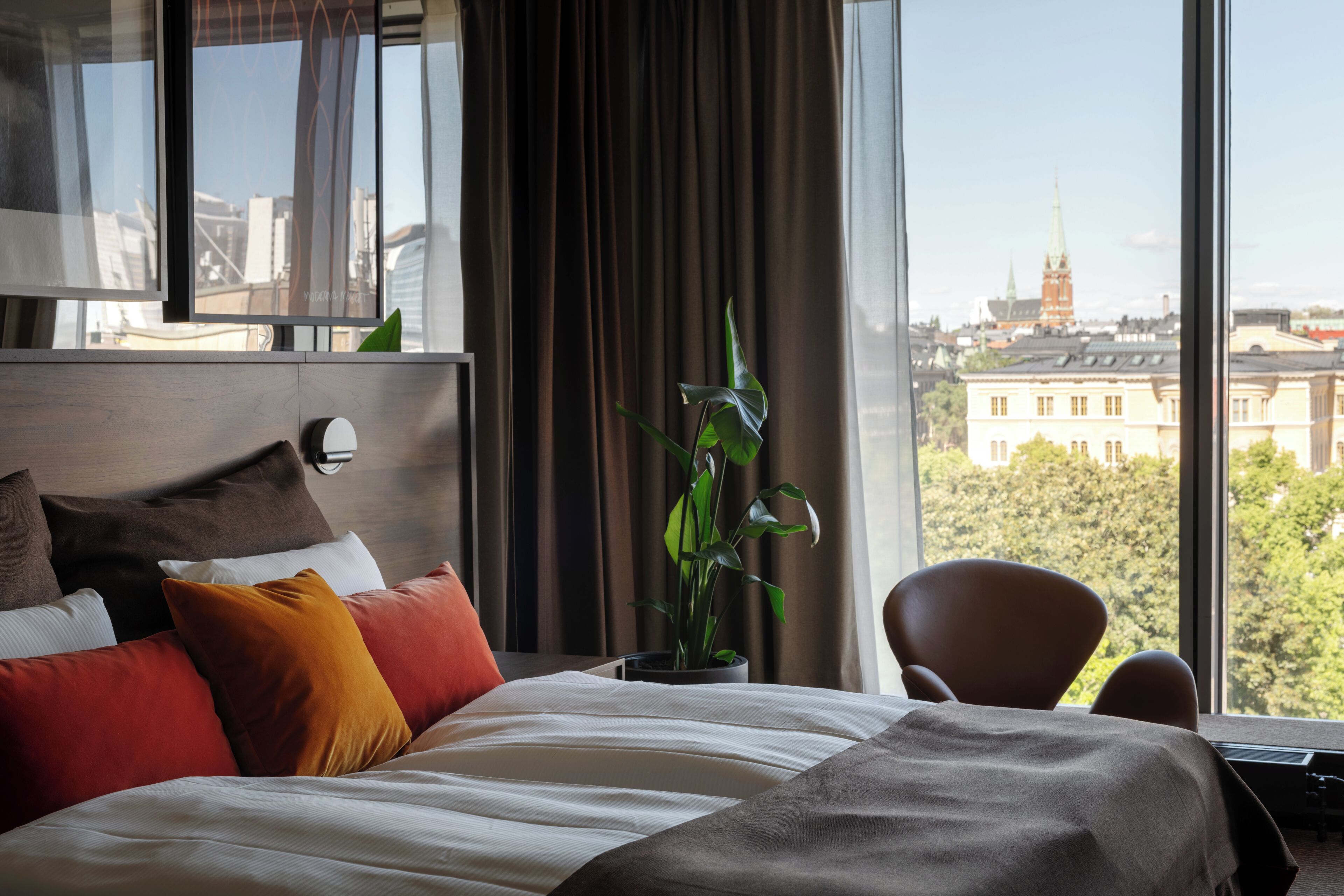 Ein Hotelzimmer mit einem großen Bett, einem Blumentopf, einem Sessel und Blick auf eine Stadtlandschaft.