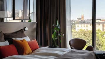Habitación de hotel con una cama grande, una planta en maceta, una silla y vistas a un paisaje urbano.