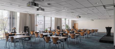 Un salón de conferencias con varias mesas y sillas, ventanas amplias y un escenario con un atril.