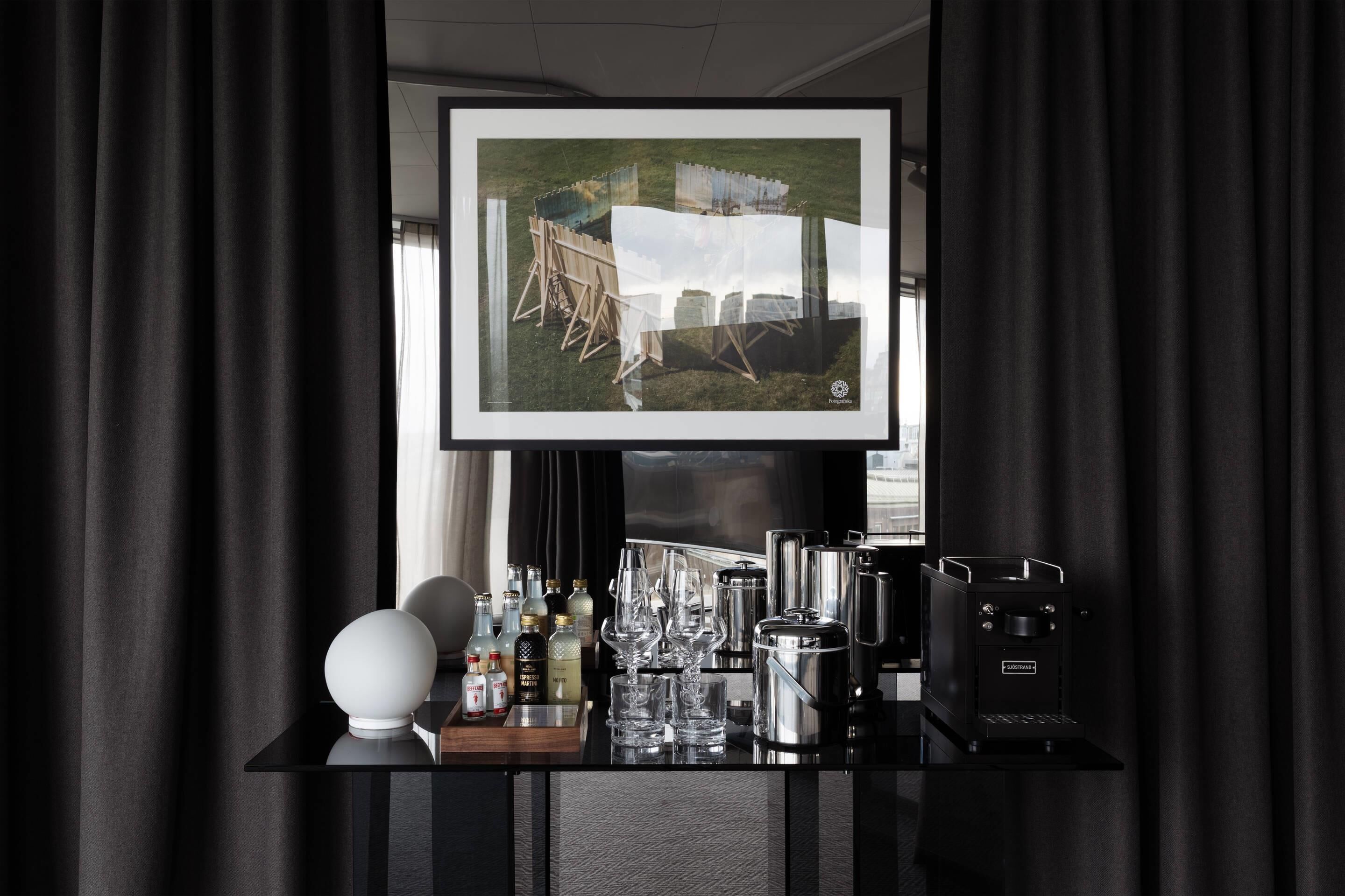 A bar area with a television displaying a landscape, a coffee machine, and various bottles and glasses on a black counter.