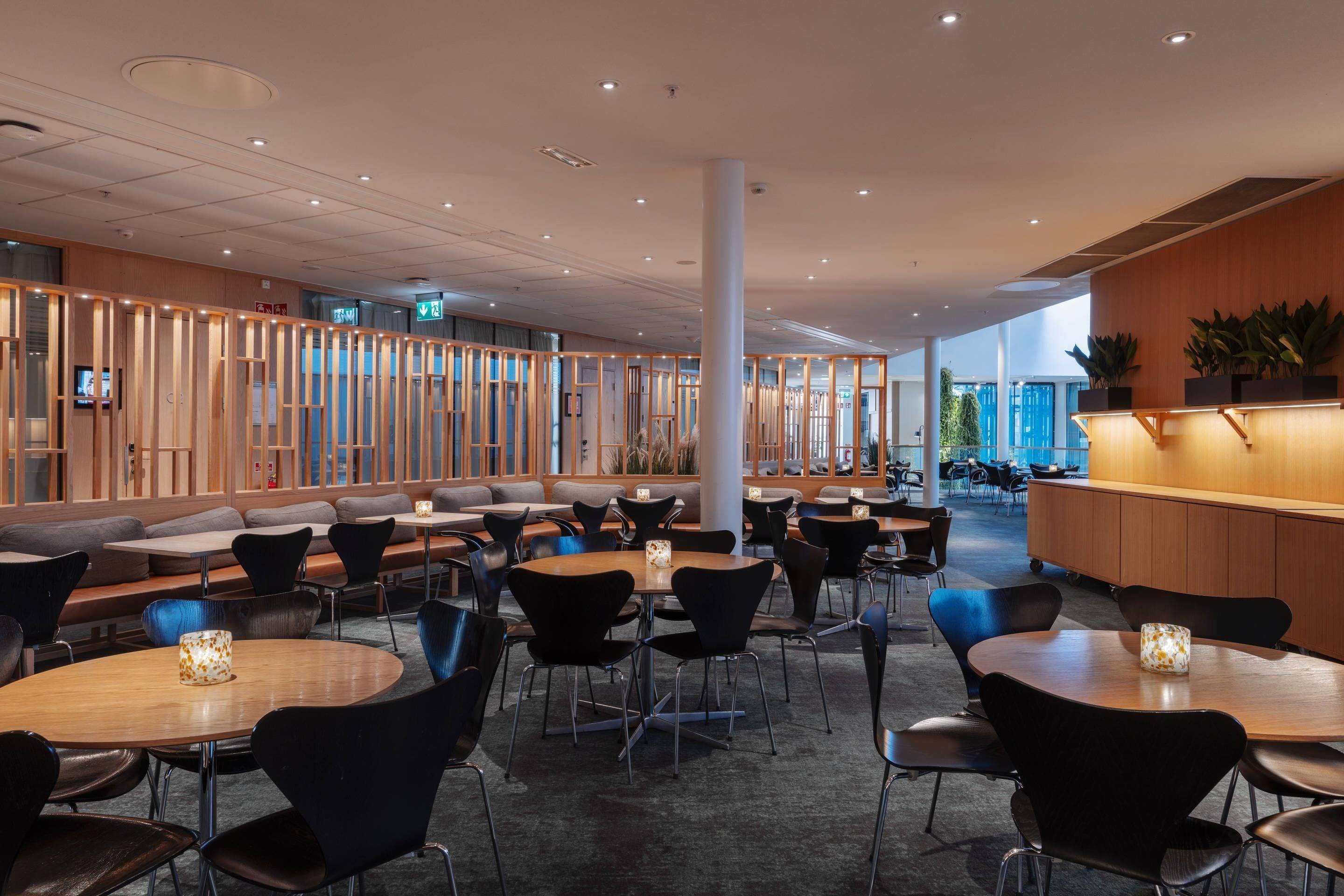 A modern indoor space with round tables and black chairs, wooden partitions, and a reception area with a counter and plants.