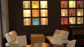Une pièce avec une table en bois et quatre chaises blanches, un mur sombre orné de panneaux carrés éclairés, et un petit meuble en bois.