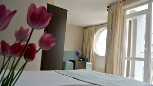 Une chambre d’hôtel avec un lit, une table de chevet, une fenêtre avec des rideaux et un vase rempli de fleurs.
