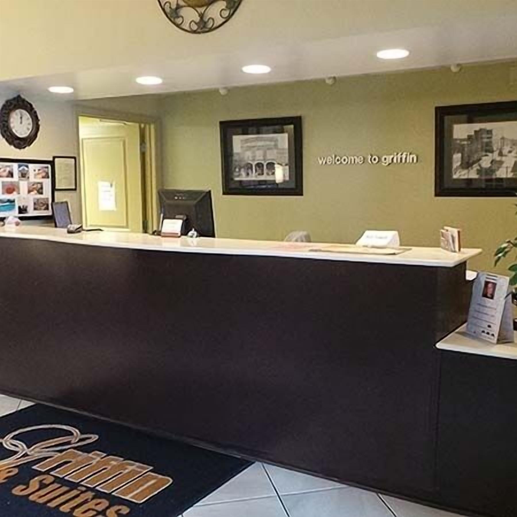 Zona de recepción de un hotel con mostrador de recepción, un reloj, cuadros en la pared y un cartel de bienvenida.