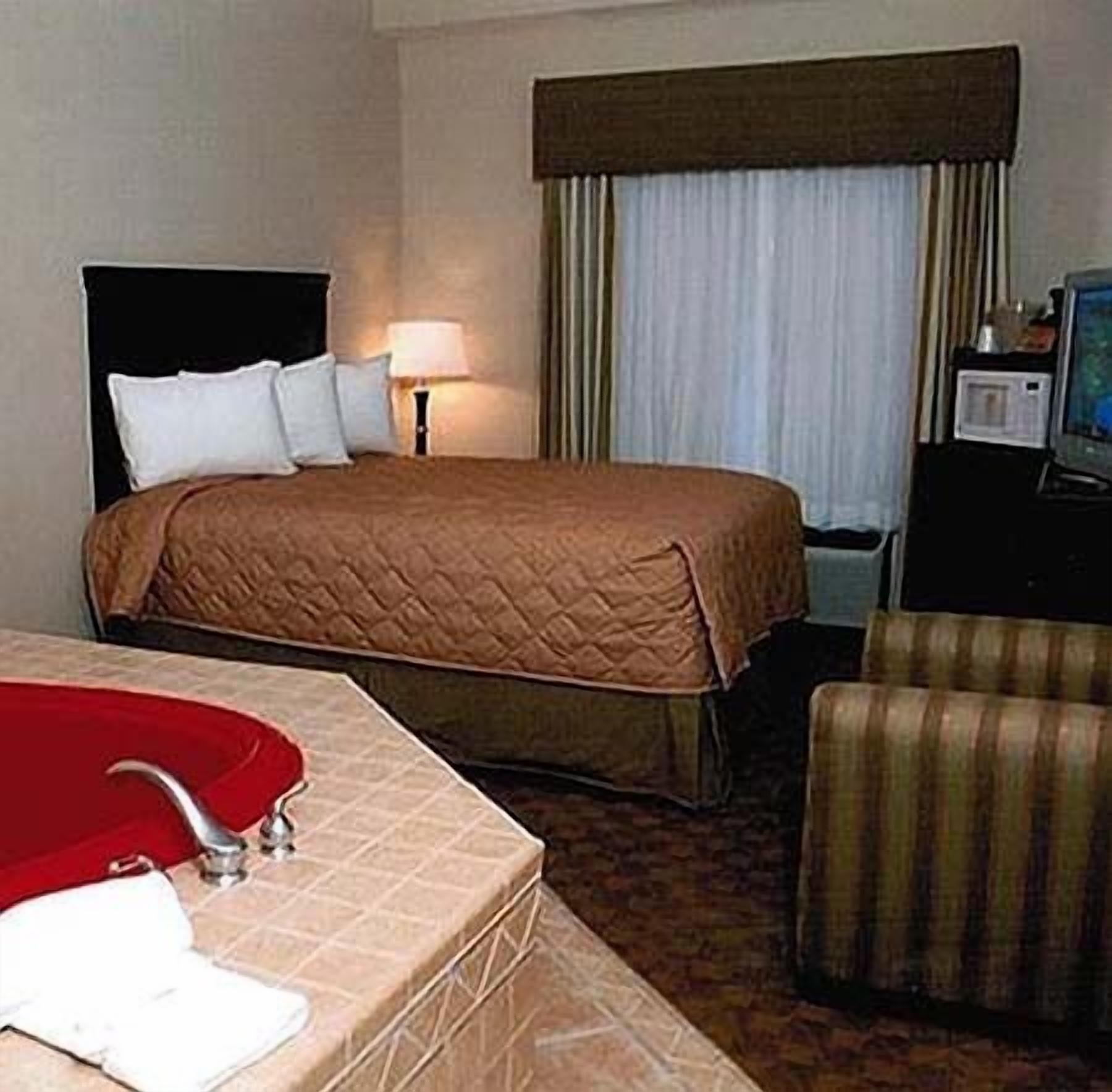 A hotel room with a bed, a bedside table with a lamp, a TV, a chair, and a bathroom with a sink and mirror.