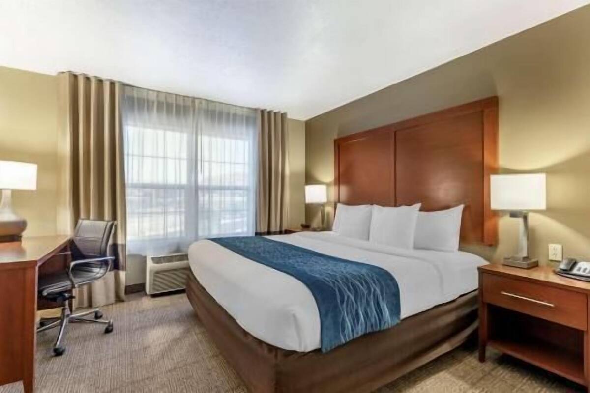 A hotel room with a large bed, a desk, a chair, a nightstand, and a window with curtains.
