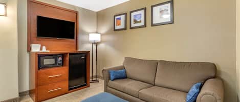 Une chambre d’hôtel équipée d’un téléviseur à écran plat fixé à un meuble en bois, d’un four à micro-ondes, d’un petit réfrigérateur, d’un canapé avec des coussins bleus et d’une lampe sur pied.