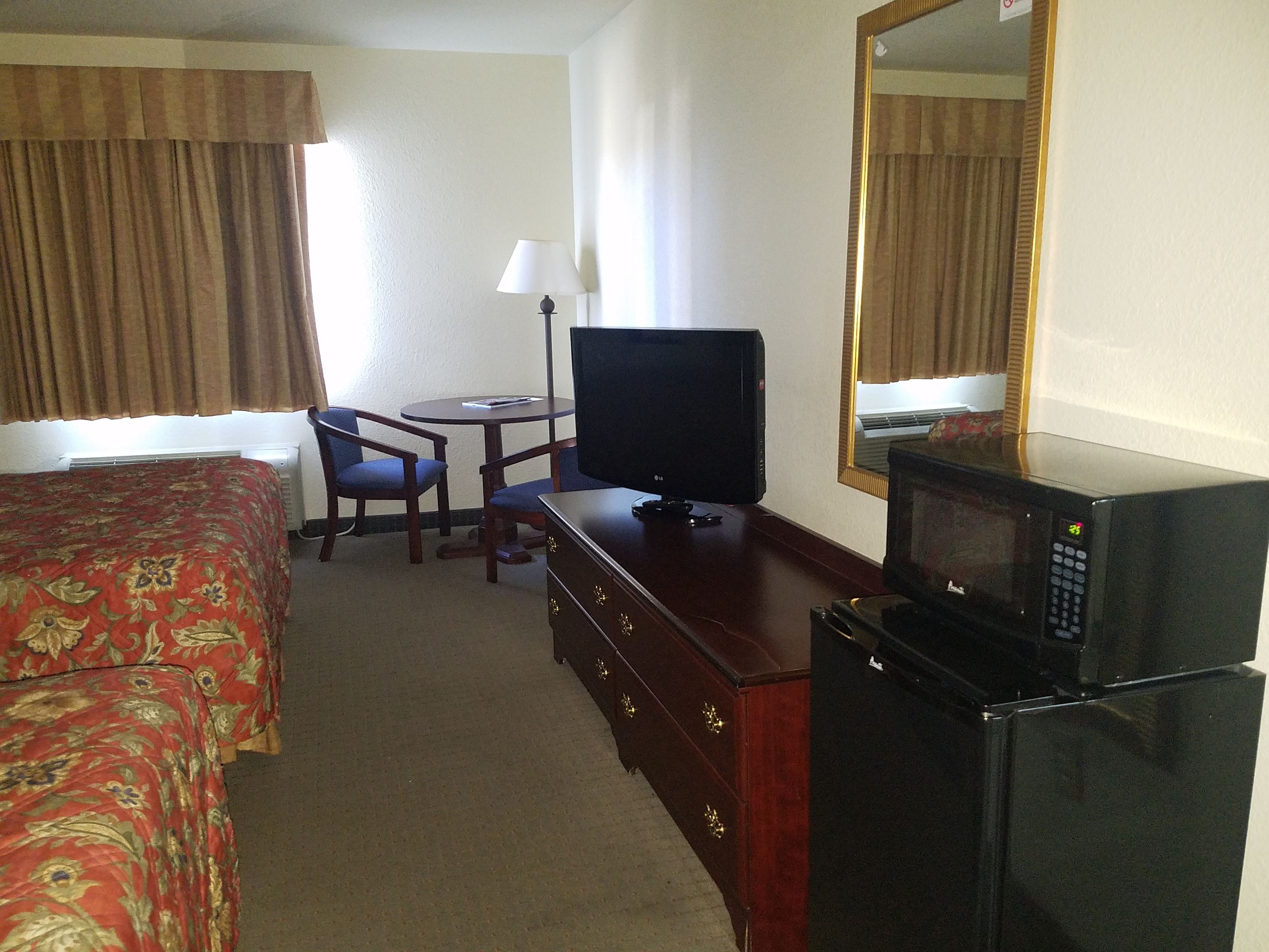 A hotel room with a bed, a desk with a television, a chair, a dresser, and a microwave.