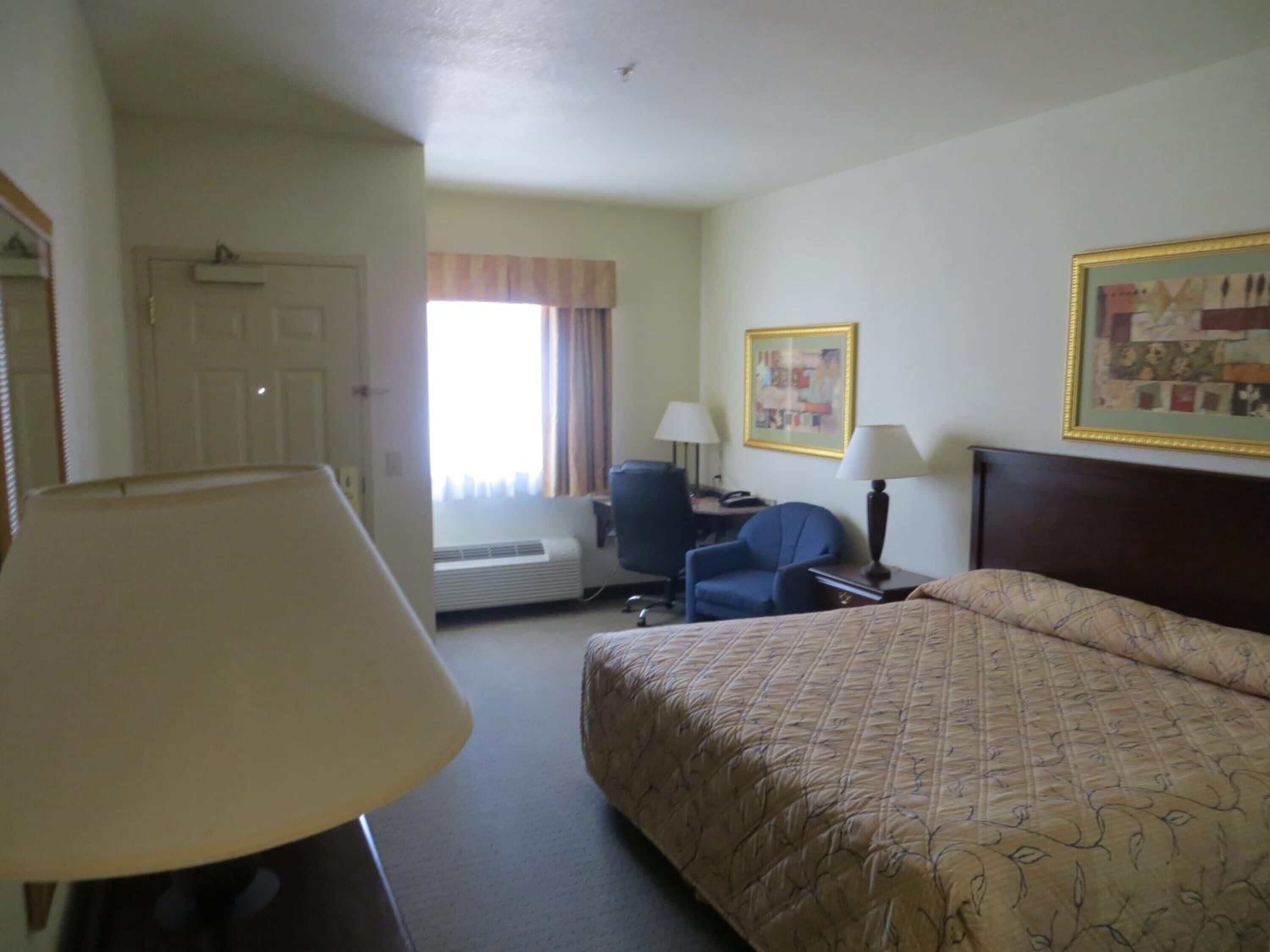 A hotel room with a bed, a desk, a chair, a lamp, a window with curtains, and a painting on the wall.