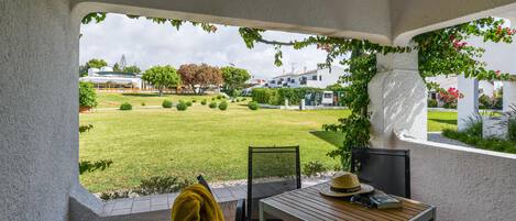 En patio med et bord og stole, et gult håndklæde og udsigt til et græsområde med træer og bygninger.