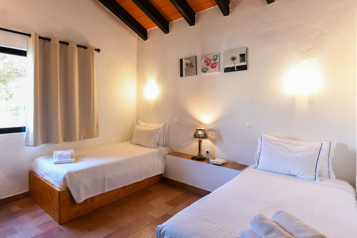 A room with two beds, a nightstand, and a lamp. There are wall-mounted lights and a window with curtains.