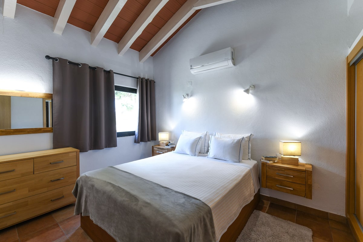 A bedroom with a bed, bedside tables, a dresser, an air conditioning unit, and a window with curtains.