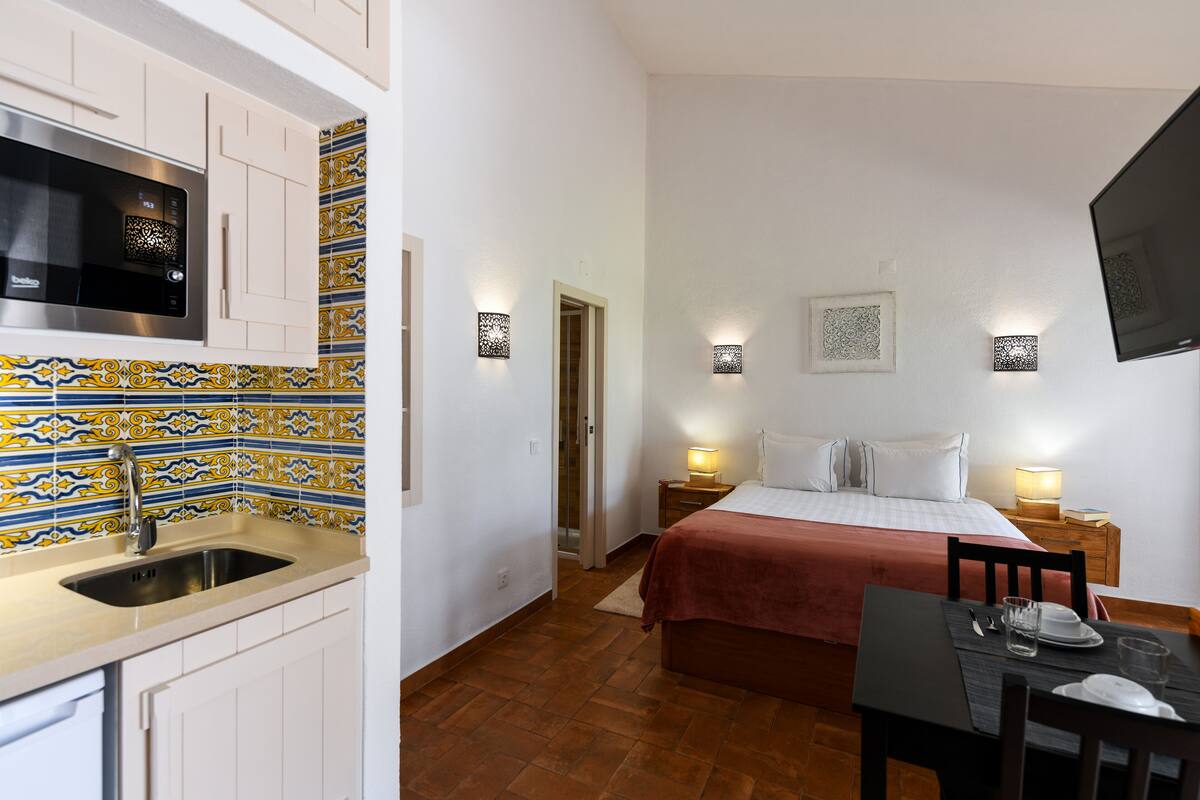 A compact hotel room with a kitchenette, a bed with a red blanket, bedside lamps, and a flat-screen TV mounted on the wall.