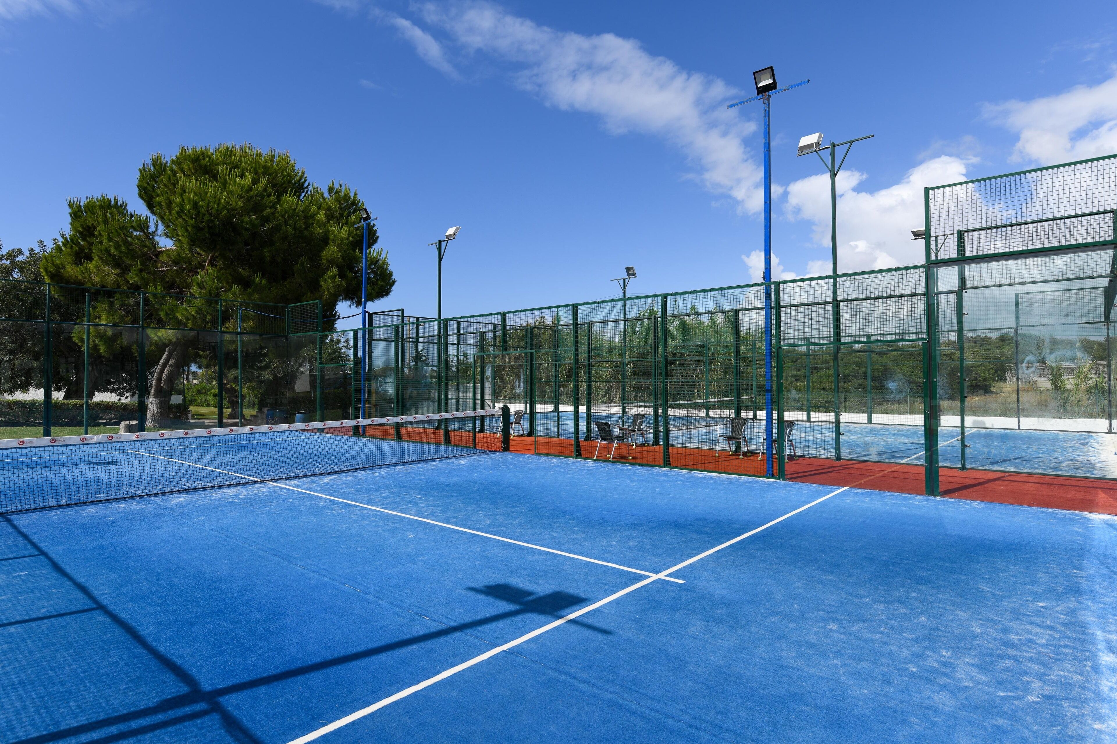 Ein Tennisplatz im Freien mit blauem Belag, umgeben von einem grünen Zaun und mit Flutlicht ausgestattet.