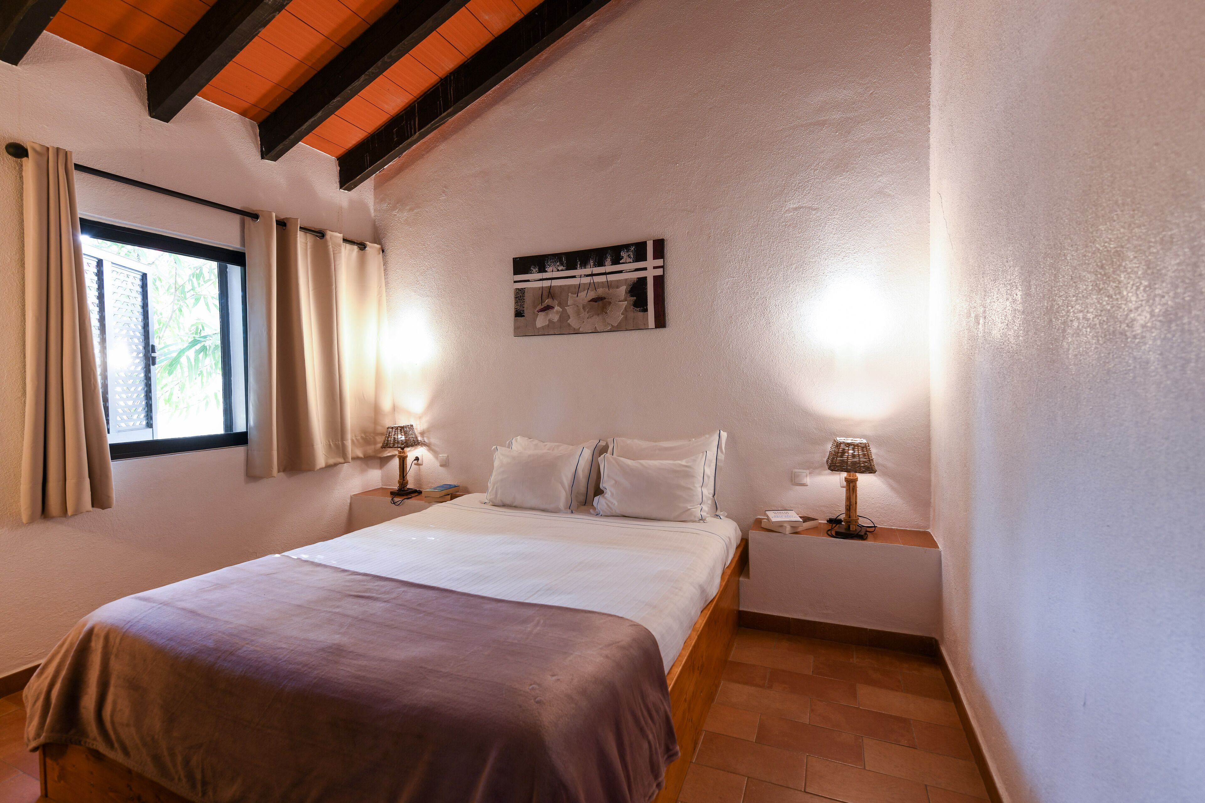 Slaapkamer met een bed, twee nachtkastjes, een raam met gordijnen, een aan de muur hangend schilderij en een houten plafondbalk.