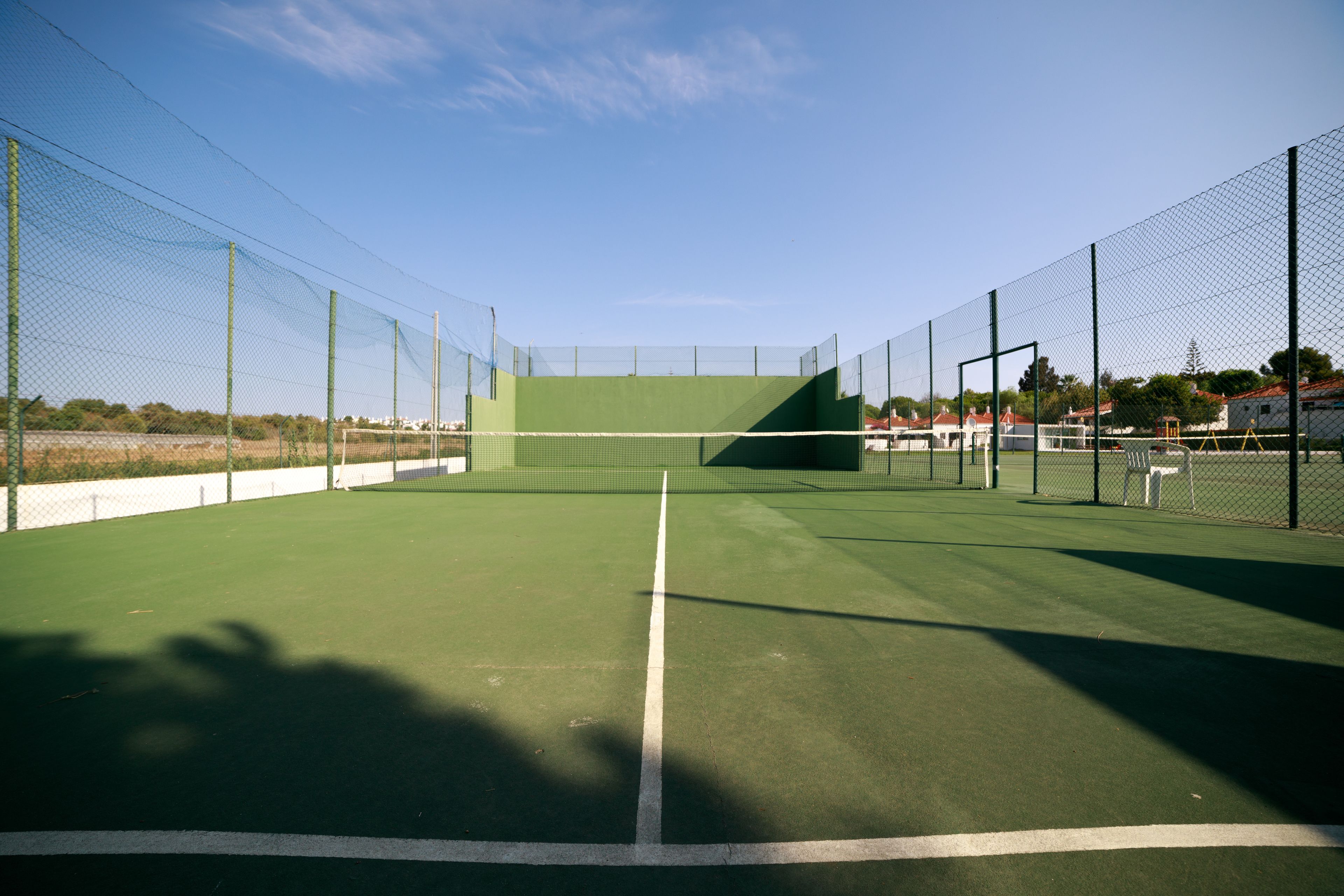 Una cancha de tenis con red, rodeada por una cerca de malla metálica, y un cielo despejado arriba.