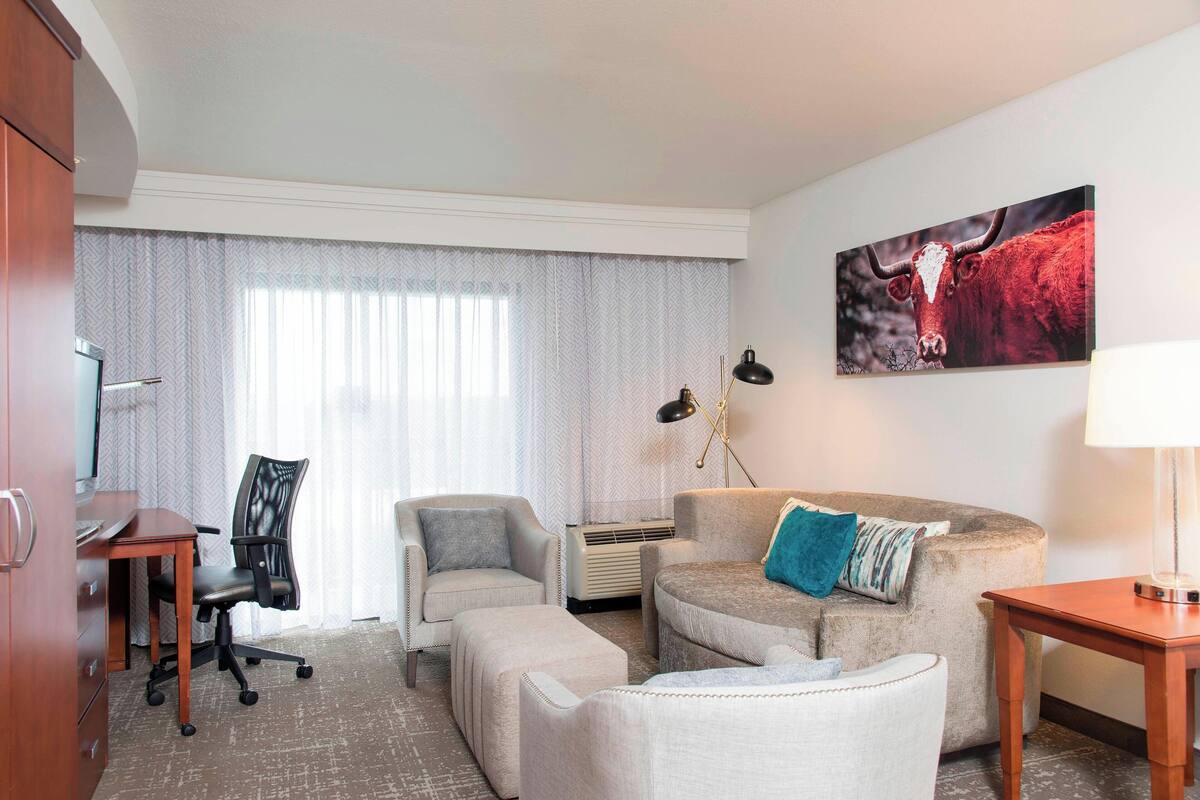 Quarto de hotel com escrivaninha, cadeira de escritório, sofá e banqueta. Possui uma janela grande com cortinas, uma luminária fixada na parede e uma pintura de um touro.