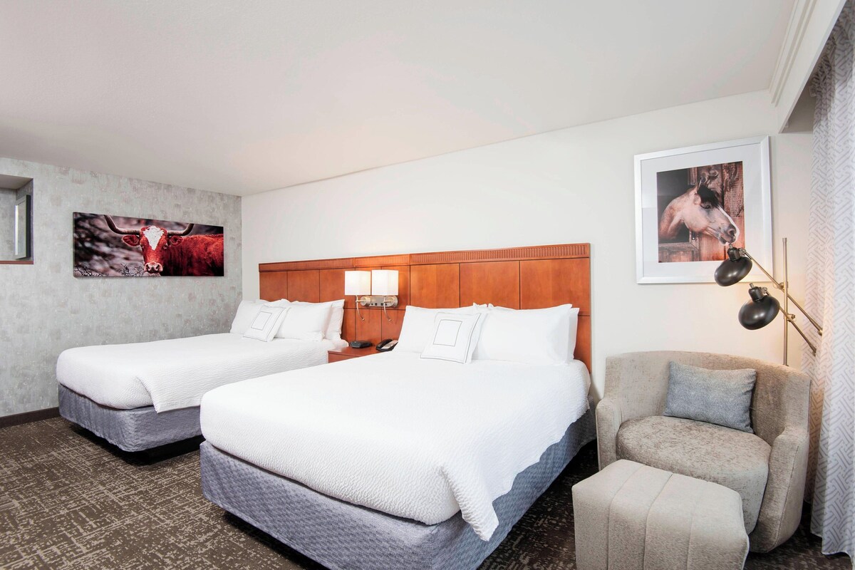 A hotel room with two beds, a wooden headboard, a desk with a lamp, and a framed picture on the wall.
