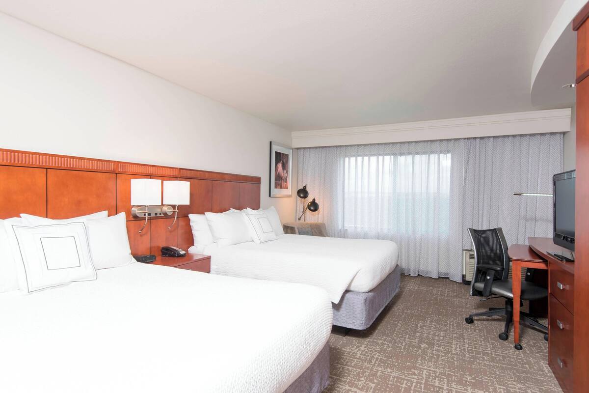 Quarto de hotel com duas camas, uma escrivaninha com cadeira, televisão e janela com cortinas.