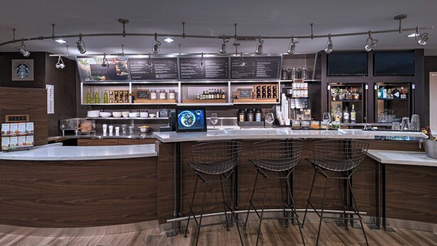 Zona de bar moderna con mostrador de madera, tres taburetes de metal y una pantalla digital con el menú.