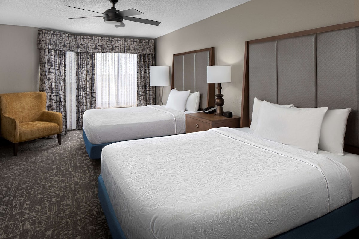 A hotel room with two beds, a chair, a nightstand with lamps, and a ceiling fan.