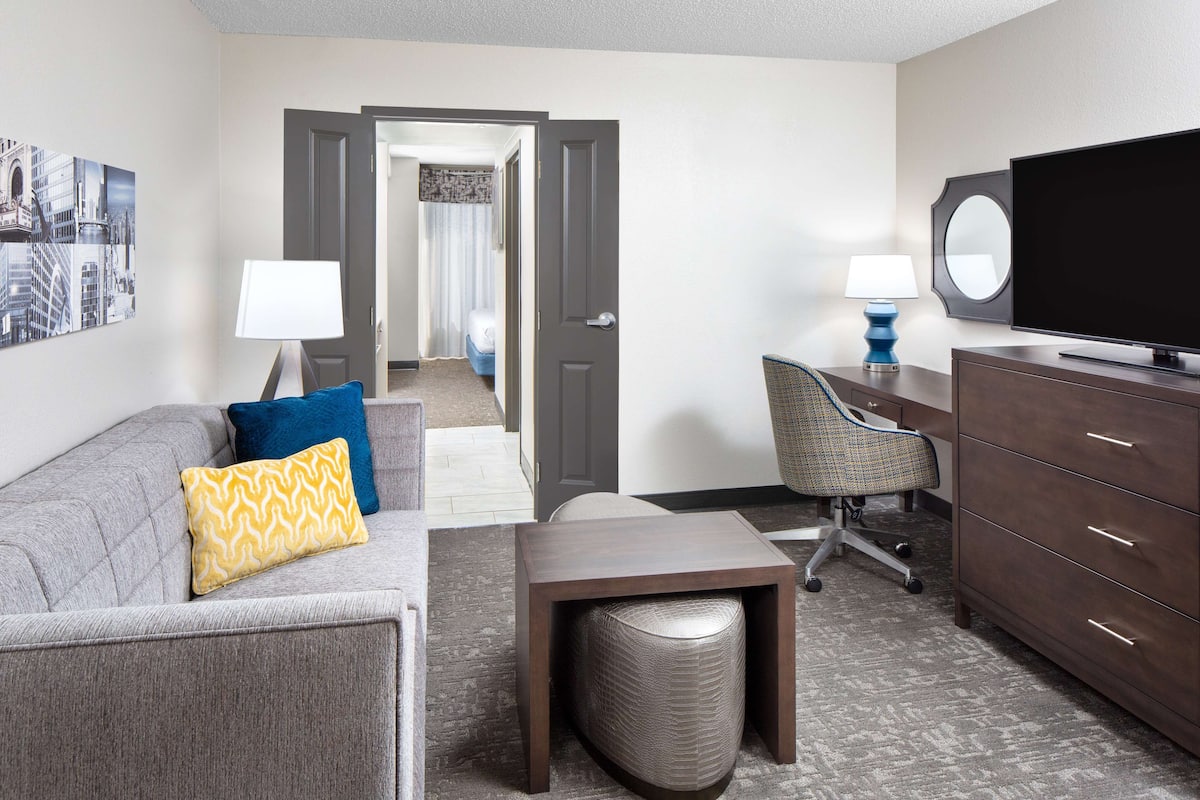 A hotel room with a grey sofa, a small table, a TV, a lamp, and a mirror.