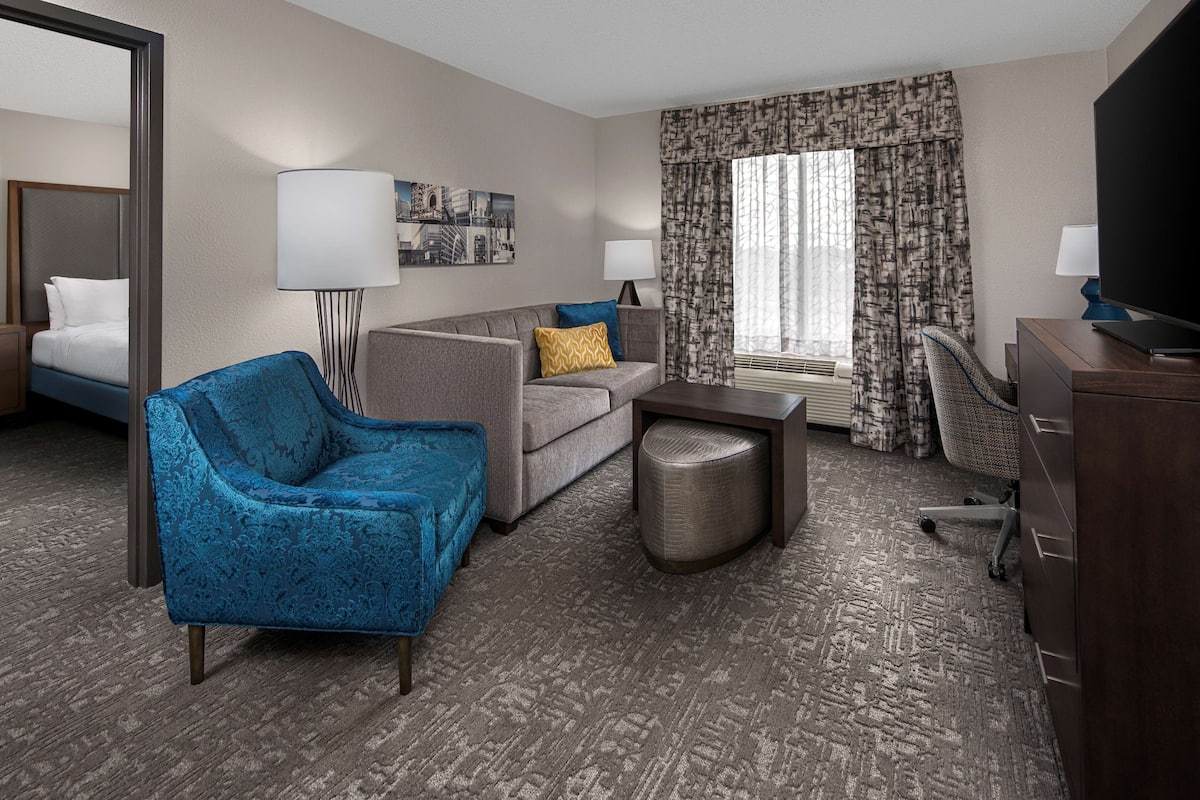 A hotel room with a bed, a blue armchair, a grey sofa, a small table, a lamp, a TV, and a window with curtains.