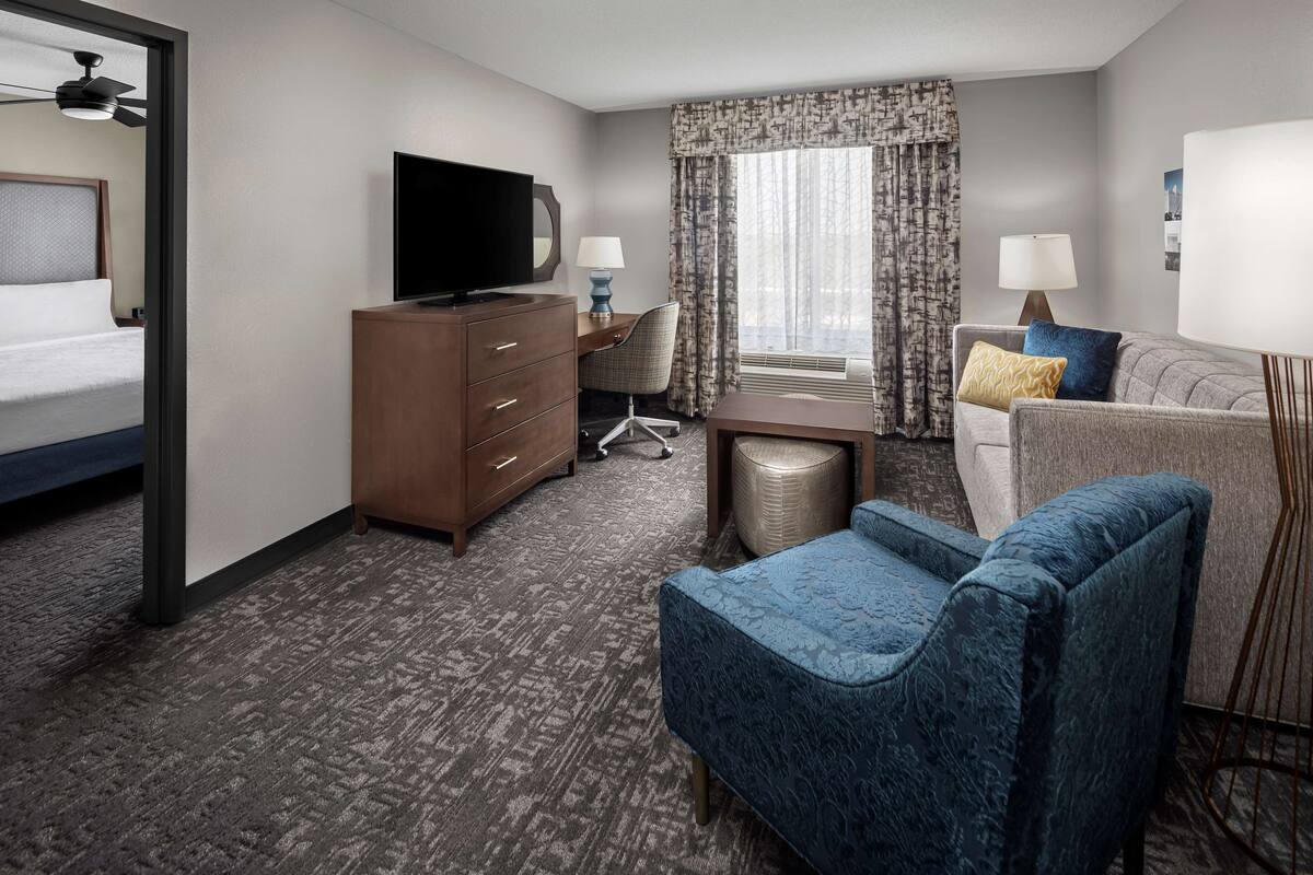 A hotel room with a bed, a desk with a chair, a TV, a sofa, and a large window with curtains.