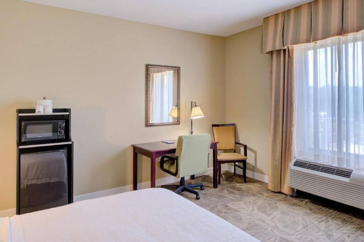 A hotel room with a bed, a desk with a chair, a microwave, a mirror, and a window with curtains.