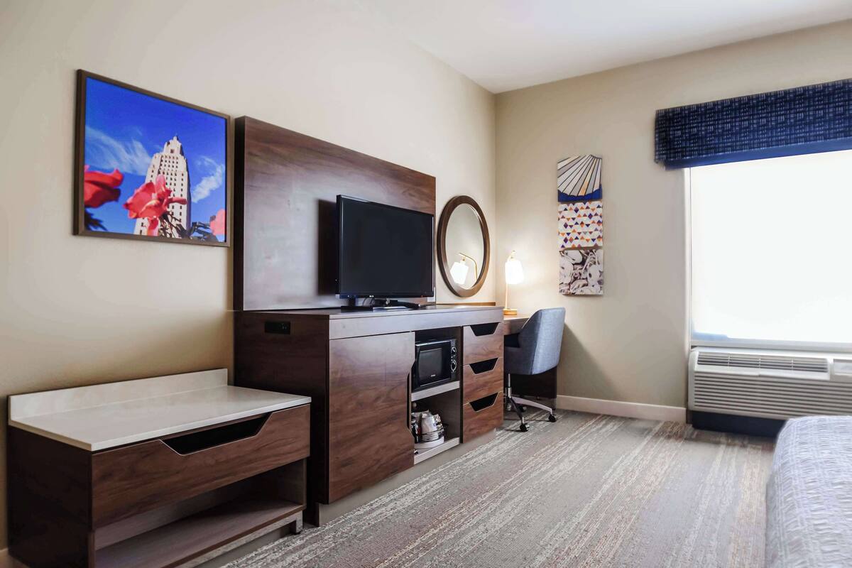 A hotel room with a wooden desk, a flat-screen TV, a chair, a bed, a window with a view, and artwork on the walls.