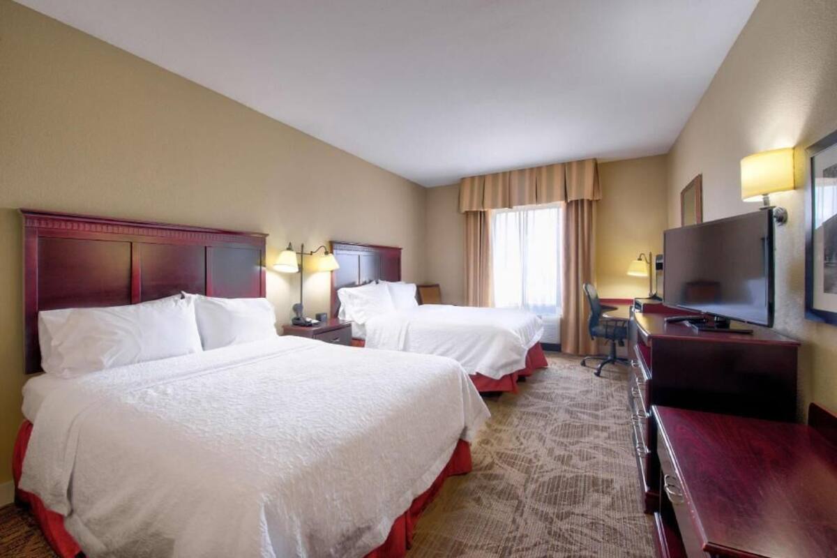A hotel room with two beds, a desk, a television, and a window with curtains.