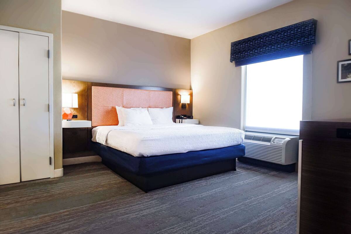 A hotel room with a large bed, a nightstand, a desk, a wardrobe, and a window with blinds.
