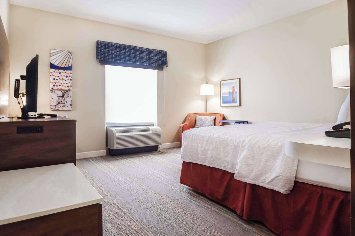 A hotel room with a bed, a desk, a chair, a TV, a lamp, and a window with a view.