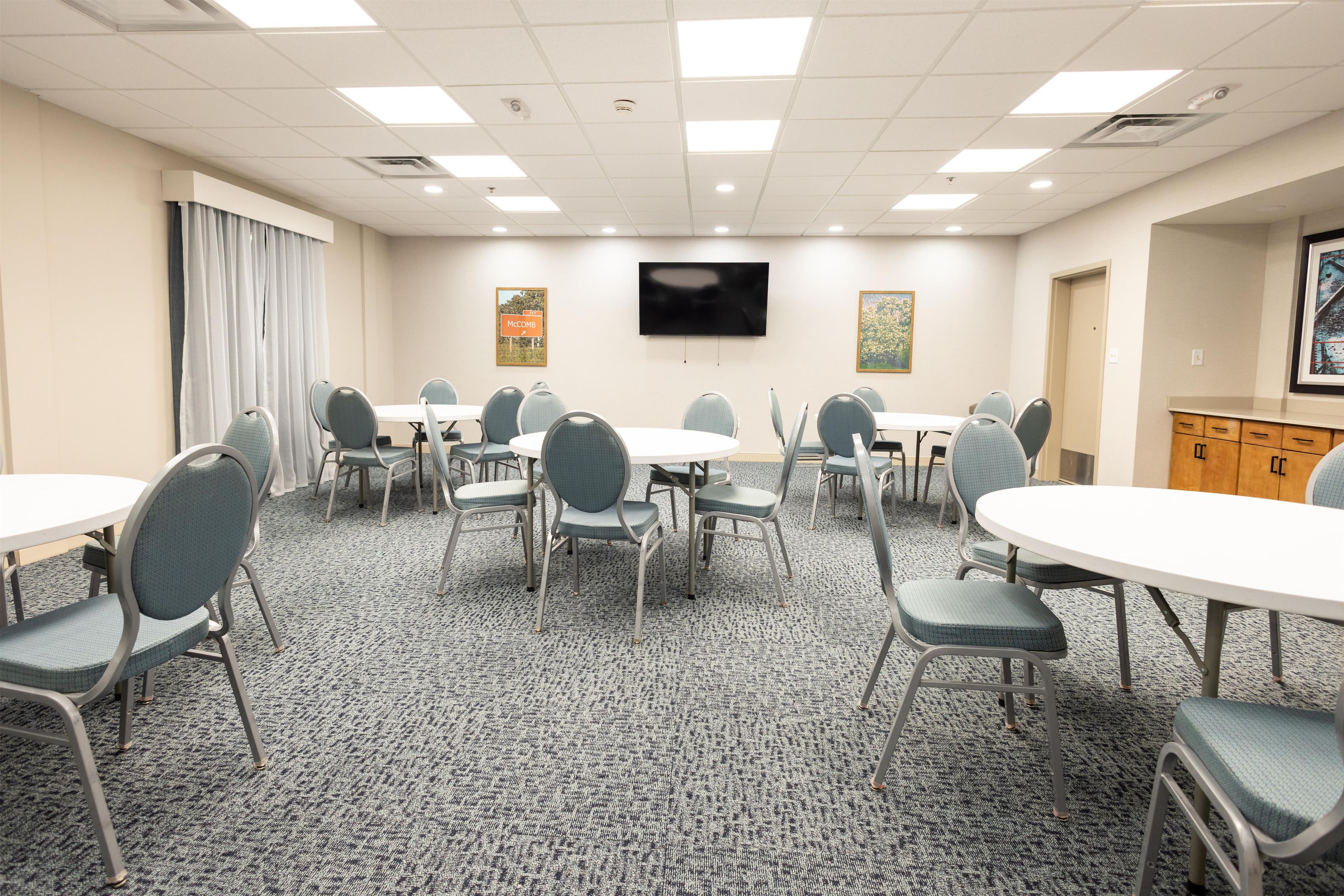 A conference room with multiple tables and chairs, a flat-screen TV mounted on the wall, and a carpeted floor.