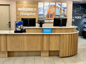 Un mostrador de recepción de hotel con un letrero que dice 'Welcome to Hampton', una insignia de 'Hilton Honors' y un letrero de 'Tastes' que indica una zona de servicio de comidas.